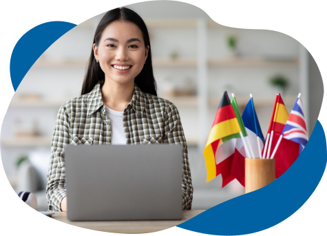 A smiling young asian woman sits at a desk with a laptop, surrounded by a cup filled with various international flags. she's wearing a plaid shirt in an office setting.