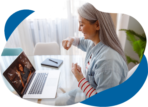 A senior woman with gray hair smiling and engaging with someone over her laptop, gesturing a fist bump towards the screen, in a brightly lit home office.