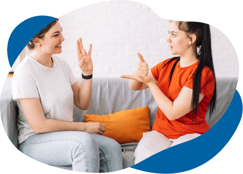 Two women sitting on a couch, engaging in conversation using sign language. one is wearing a white shirt and blue headband, the other in a red shirt. they smile and gesture animatedly.